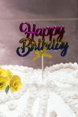White heart shaped birthday cake. Decorated with yellow flowers and white whipped cream with the inscription Happy Birthday