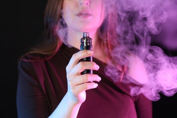 Woman using electronic smoking device in neon lights on black background, closeup