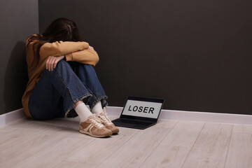 Cyber bullying. Upset teenage girl and laptop with text You Loser on floor indoors. Space for text