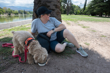 Woman relaxes by a lake under a tree while her dog drinks water from a bowl during a sunny park outing.