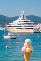 Close-up of pastel-colored ice cream cone in soft blue, pink, and yellow tones, with a luxury yacht blurred in the background, seaside vacation vibe