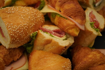 Assorted sandwiches with sesame rolls and croissants, filled with cheese, lettuce, and deli meats like salami or ham, shown in a close-up view.