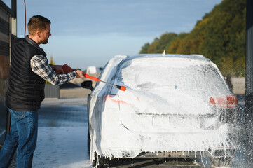 A man washes off the foam while washing the car. A vehicle is engulfed in soapy foam during a car...