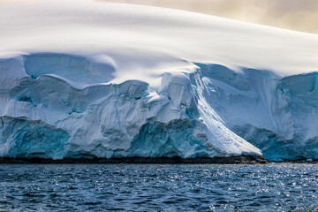 Snowy shoreline of Antarctic Peninsula. Blue ocean in foreground; gray clouds in background....