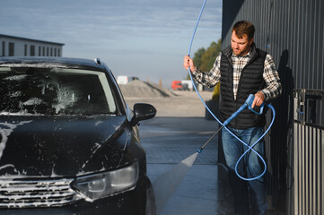 A man washes his car at a self service car wash using a hose with pressurized water.