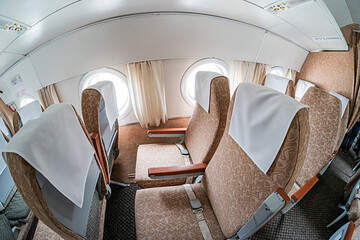 Empty airplane cabin interior, multiple rows of beige-covered seats, carpeted floor, overhead bins,...