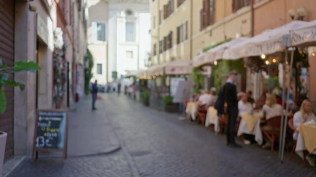 Blurred outdoor cafe scene in rome with bokeh effect showing pedestrian street and people dining under umbrellas along a historic cobblestone street.