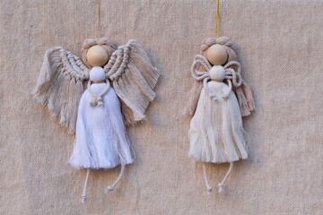 Two handmade macrame angels in neutral tones, featuring wooden heads and textured cords. Hanging against natural fabric background. Ideal for rustic, Christmas decor, DIY crafts, and natural themes