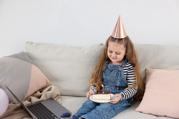 Birthday celebration. Little girl with piece of cake having video call via laptop on sofa at home