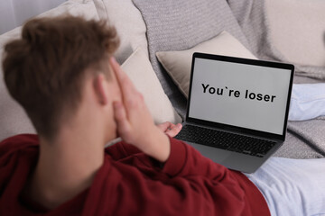 Cyber bullying. Upset teenage boy looking at laptop with text You Loser on sofa at home, closeup