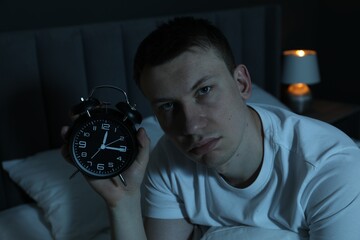 Frustrated man with alarm clock suffering from insomnia on bed indoors