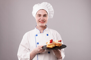 Confectioner in uniform holding delicious profiteroles on light grey background