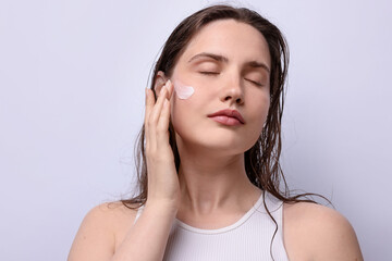 Beautiful woman applying skincare product onto her face on light grey background, closeup