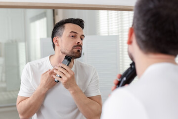 Handsome man shaving with trimmer near mirror at home
