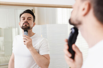 Handsome man shaving with trimmer near mirror at home