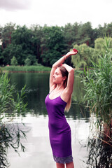 Young blonde woman in dress standing near lake during summer sunset. Young woman posing on camera...