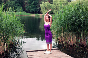Young blonde woman in dress standing near lake during summer sunset. Young woman posing on camera...