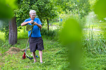 Senior man maintaining grass trimmer in backyard. An older man wearing protective goggles cleans...