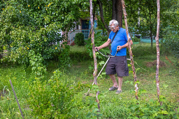 Older man trimming backyard grass near garden plants. Senior man uses a string trimmer to cut...