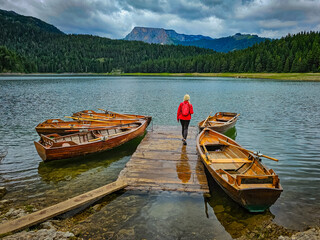 A girl in red walks over a platform with wooden boats on the lake - Black Lake, Montenegro