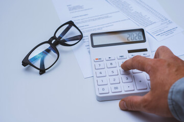 White calculator displaying the number 2026, placed on financial documents with black eyeglasses nearby. Concept business planning and tax calculation.