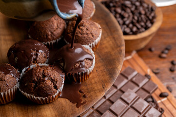 Chocolate muffins with liquid chocolate and dulce de leche with cocoa powder, a chocolate bar, and coffee beans on a wooden table with kitchen utensils.