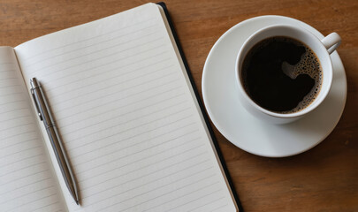 Open notebook with lined paper, pen sits on wooden table beside cup of dark coffee. Notebook blank, ready for writing notes journaling. Coffee cup full with crema, suggesting morning work study