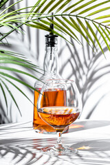 Elegant glass of amber cognac beside a bottle against tropical foliage casting shadows in a stylish...