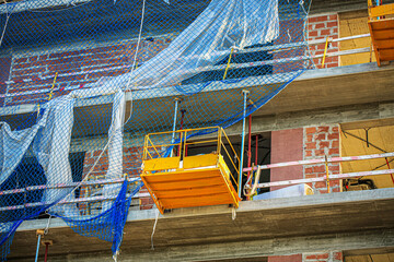 Construction site detail with safety netting, suspended lift platform, and exposed red brick walls....