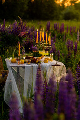Romantic table decor for a loving couple on the blooming meadow with purple lupines. Two glasses of limoncello, flowers in a vase, candles, lemons, wooden furniture. Sunset, golden hour.