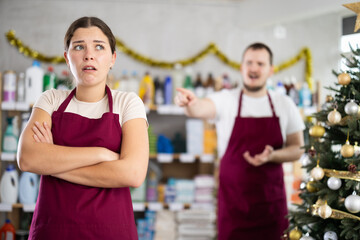 Upset young woman in maroon apron standing with arms crossed while male colleague in background...