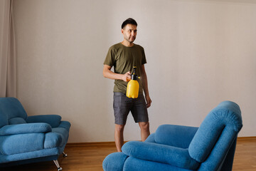 Portrait of young man in casual clothes spraying water on blue sofa using yellow plastic sprayer in...