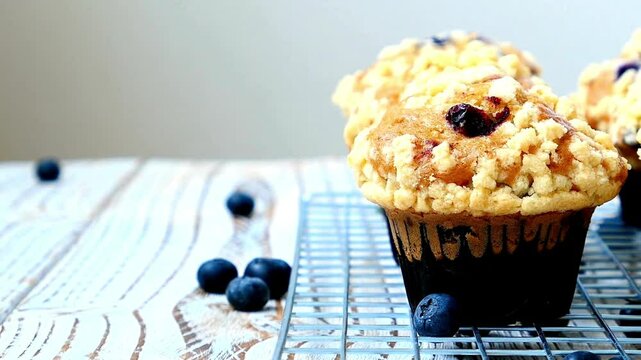 A blueberry muffin sits on a wire rack with scattered blueberries