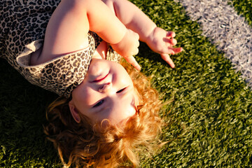A young child with curly red hair is lying on green grass smiling and playing happily under the...
