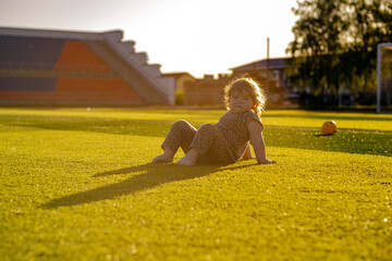 A young child sits on lush green turf during the golden hour The sun casts long shadows while the...