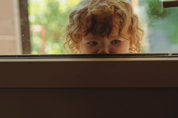 A young child with curly hair looks through a window displaying a sense of curiosity while sunlight...