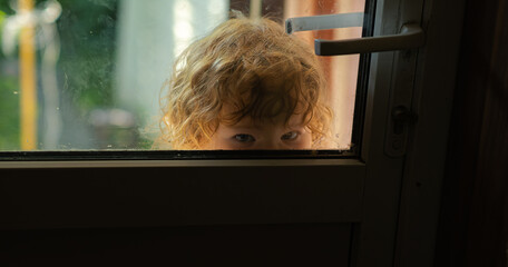 A young child with curly hair looks through a window displaying a sense of curiosity while sunlight...