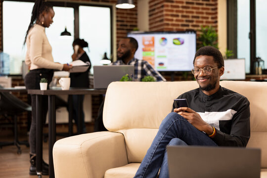 Black male manager seated comfortably on couch, using his mobile phone, taking a break from company activities. Businessman on cozy sofa, holds cellphone, checking his social media in startup office. - Powered by Adobe