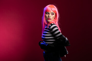 Female model with vibrant hair shows strong character in striped clothing while posing against isolated background. Pretty lady with pink wig presents clean confident stance inside fashion studio.