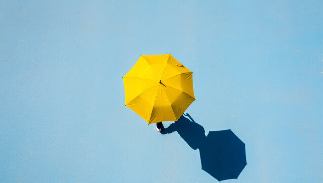 A person walks under a bright yellow umbrella casting a long shadow on a clear blue surface.