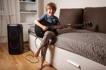 Happy boy playing electric guitar in his bedroom, sitting on the bed, laughing and enjoying music through a big speaker. Fun and creativity. Little rock star playing guitar.