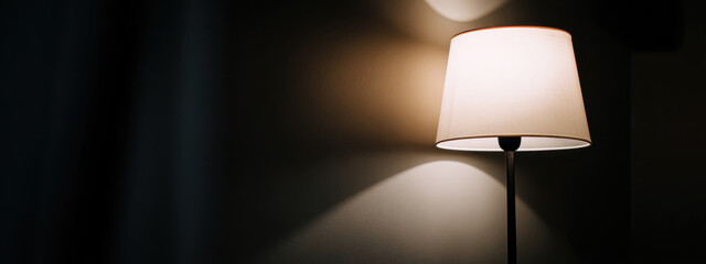 A floor lamp turned on near a wall in a dark room. Warm light creates soft shadows and a cozy ambiance. Minimalist interior in Scandinavian style.