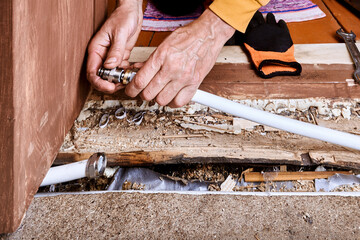 Person connects compression fitting to white metal composite pipe near damaged wooden floor.