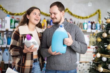 Happy young couple, warmly dressed, holding detergent bottles in decorated household goods store,...
