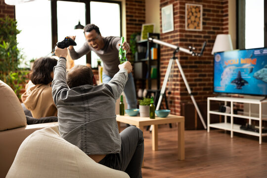 Multiethnic friends cheering and celebrating video game win during lively indoor hangout. Young diverse adults with snacks and drinks, enjoying excitement of gaming in brick wall lounging space.