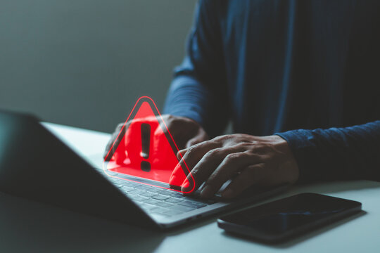Person using laptop with red warning alert triangle icon indicating cybersecurity threat, system error, data breach. online security, digital risk, hacker attack, cyber crime protection technology.