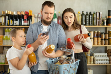 Young bearded man with wife and tween son buying organic bottled juices in grocery shop, scanning...