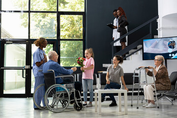 Young child offering flowers to grandfather wheelchair user during patient visit, showing...