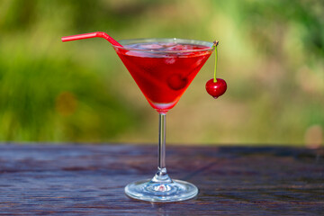 Bright red cherry cocktail in a martini glass against summer garden background on wooden table, closeup. Delicious cold alcoholic drink for relaxation on a sunny warm day