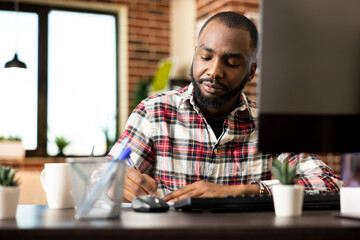 African american marketing specialist seated at desk in home office, remotely preparing startup project presentation. Focused professional writing business strategy ideas on notepad beside desktop pc.